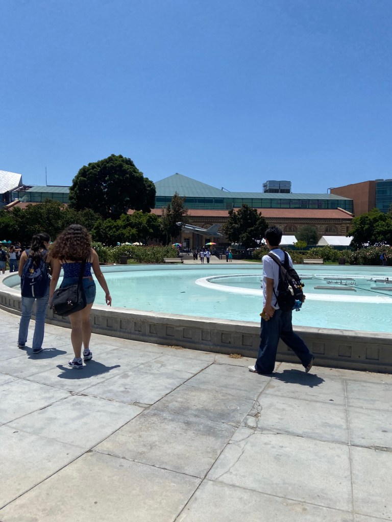 A group of three people walking beside a circular fountain in a sunny outdoor setting, with a modern building and lush greenery in the background.